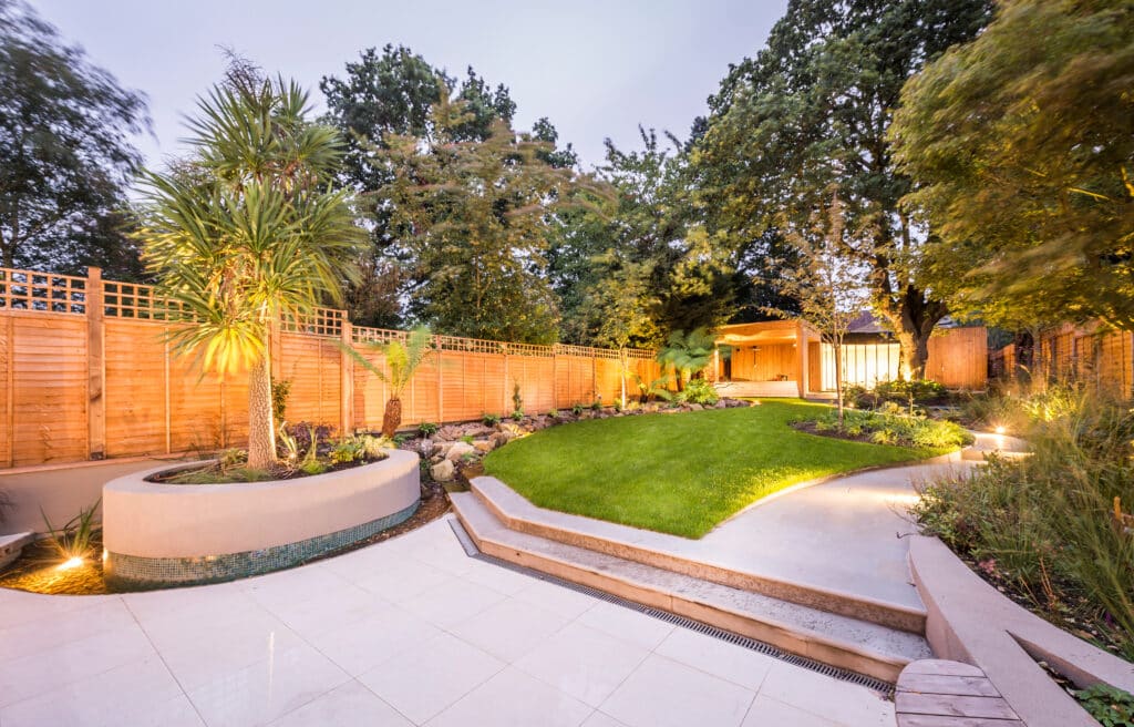 Jardin Moderne Éclairé : Terrasse et Paysage Nocturne Aménagement de jardin contemporain éclairé au crépuscule. Grande terrasse, escaliers, pelouse, clôtures en bois et palmier exotique en pot.
