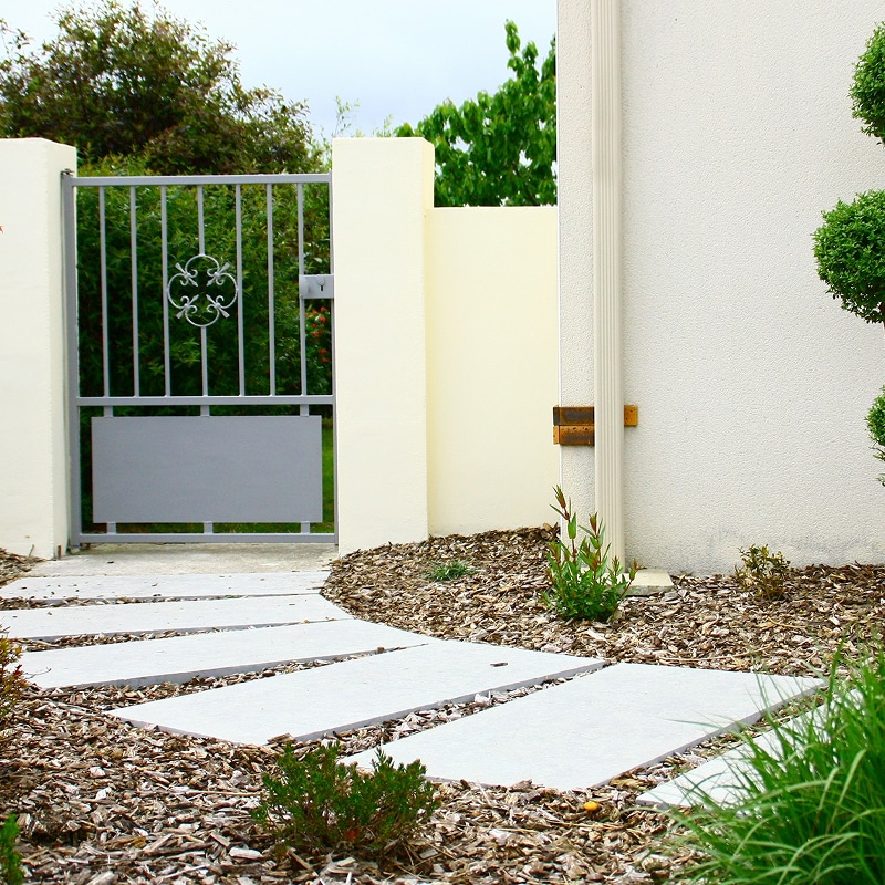 Allée de jardin moderne avec pas japonais rectangulaires sur paillis, courbant vers un portail gris en métal et des murs clairs.