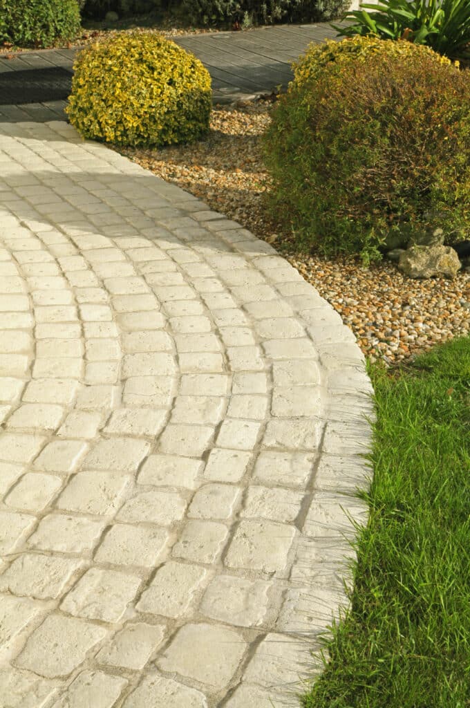 Allée de jardin courbe en pavés : Idées d'aménagement paysager. Allée de jardin courbe en faux pavés clairs, bordée de gazon vert, de gravier et d'arbustes touffus sous la lumière du soleil.