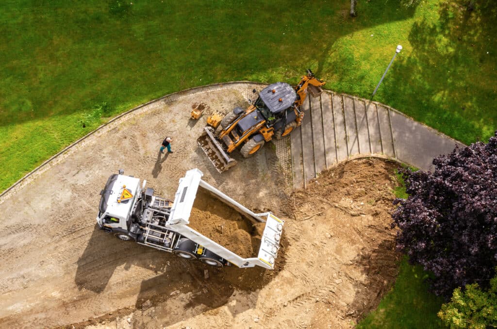 Vue aérienne des travaux. Tractopelle chargeant de la terre dans un camion-benne blanc. Ouvrier supervisant le terrassement près de la pelouse.