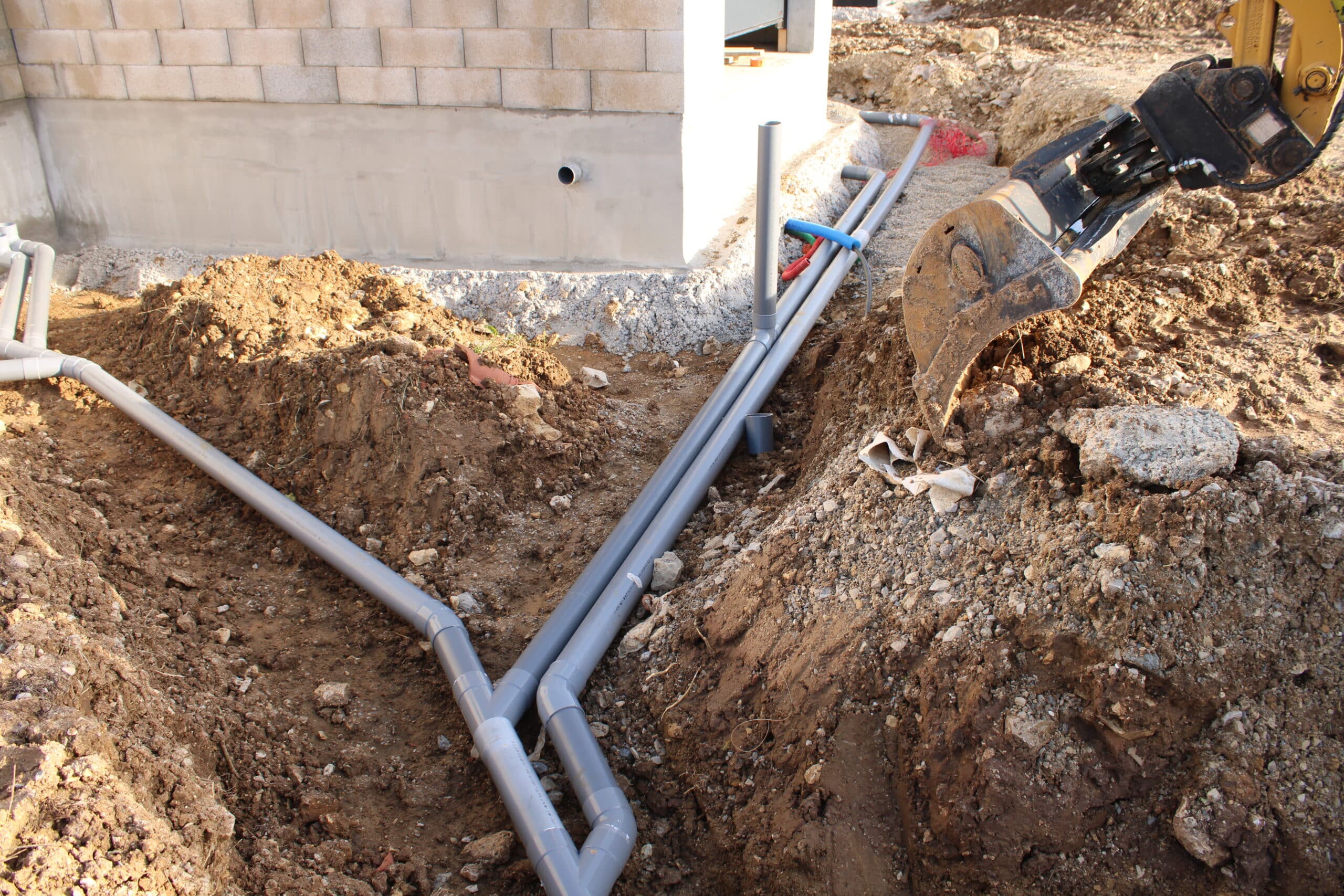 Tranchée ouverte sur un chantier de construction montrant l'installation de tuyaux PVC de drainage près d'une fondation en blocs. L'excavatrice est visible à droite.