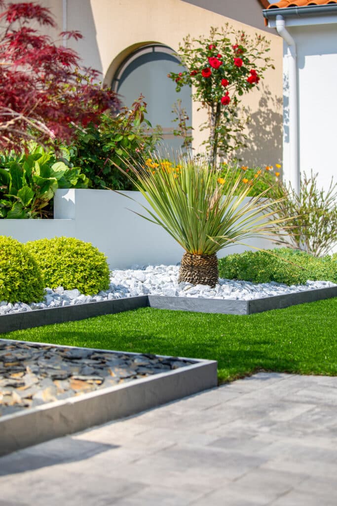 Jardin moderne design: gazon vert, gravier blanc et bordures ardoise. Aménagement paysager moderne. Pelouse artificielle verte, bordures en ardoise, yucca sur gravier blanc, et buissons topiaires. Rosier rouge contre mur beige.