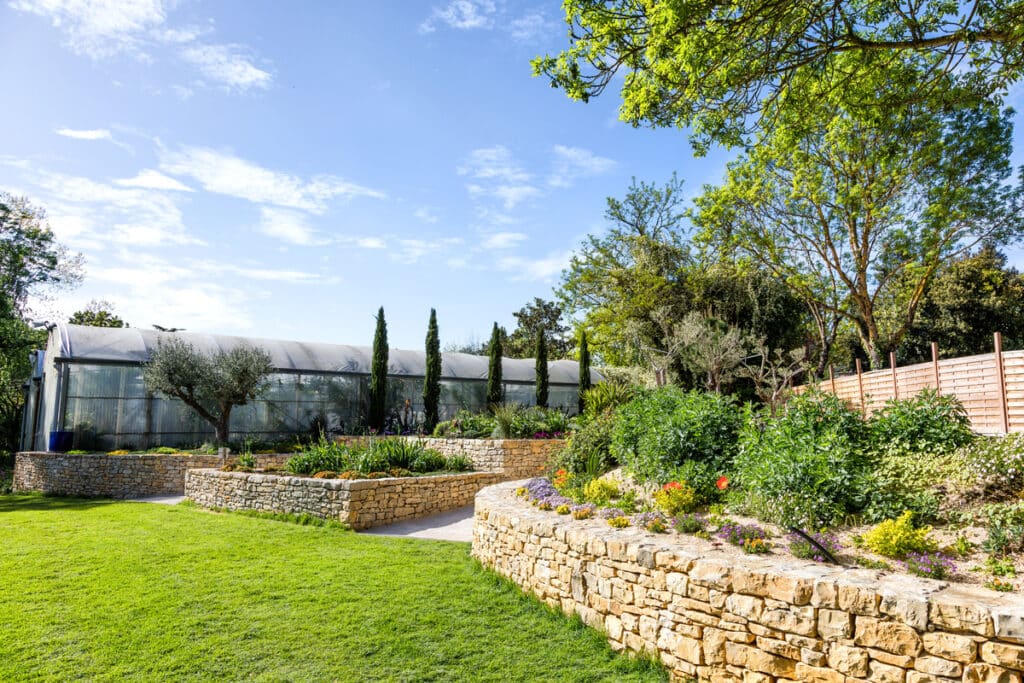Aménagement paysager : Serre et terrasses en pierre. Jardin terrassé avec murs en pierre sèche, pelouse éclatante et grands cyprès. Serre professionnelle et olivier sous ciel bleu.