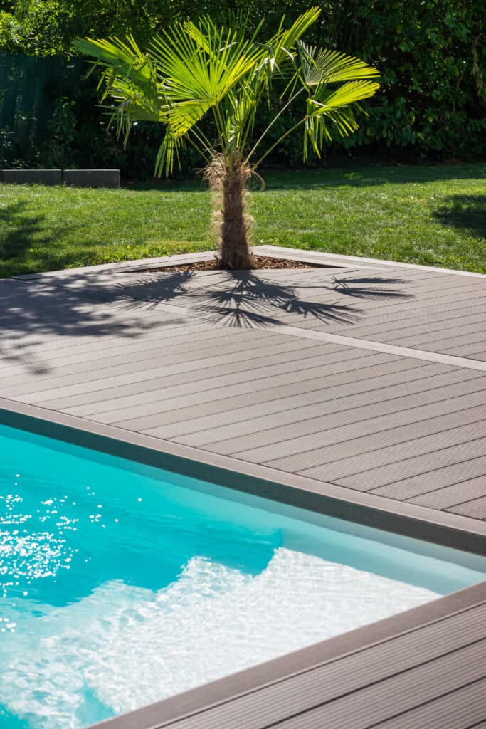 Aménagement piscine : Terrasse composite et palmier exotique. Vue en contre-plongée d'une piscine bordée d'une terrasse en composite grise. Un palmier projette ses ombres sur le pont. Eau turquoise, jardin luxuriant.
