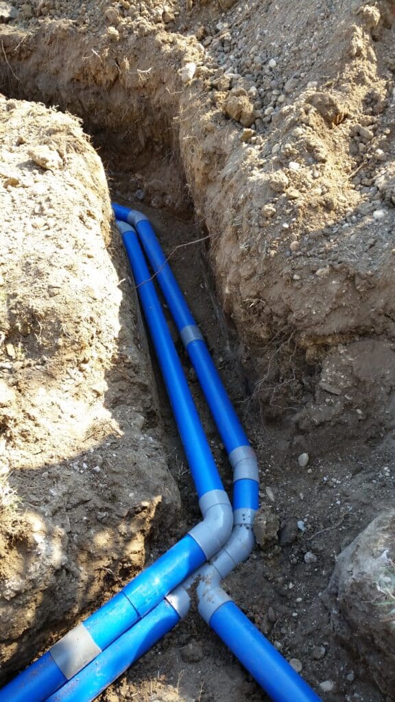 Installation de tuyaux PVC bleus dans une tranchée Deux tuyaux PVC bleus avec raccords gris installés dans une tranchée de terre pour des canalisations souterraines.