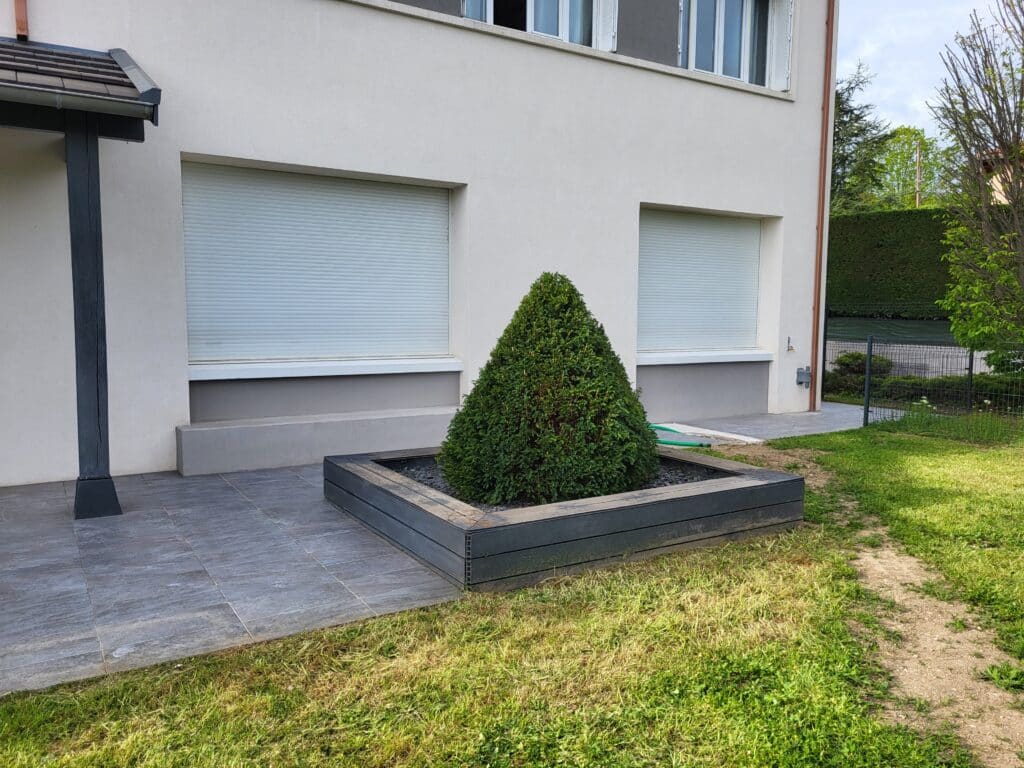 Arbuste conique en jardinière grise devant une maison moderne aux volets roulants blancs fermés.