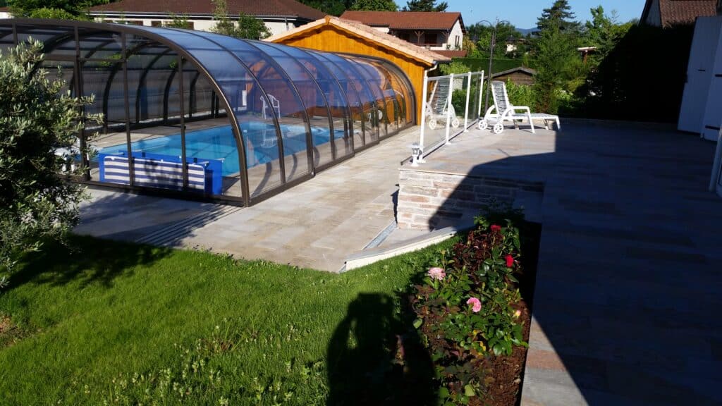 Piscine avec Abri Télescopique et Terrasse en Pierre Piscine sous abri télescopique avec grande terrasse en pierre, jardin arboré et fleurs roses.